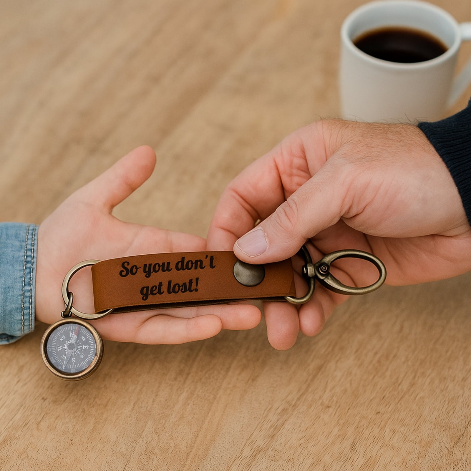 Keychain with compass and engraved text held by two hands on a wooden surface with a cup of coffee in the background.