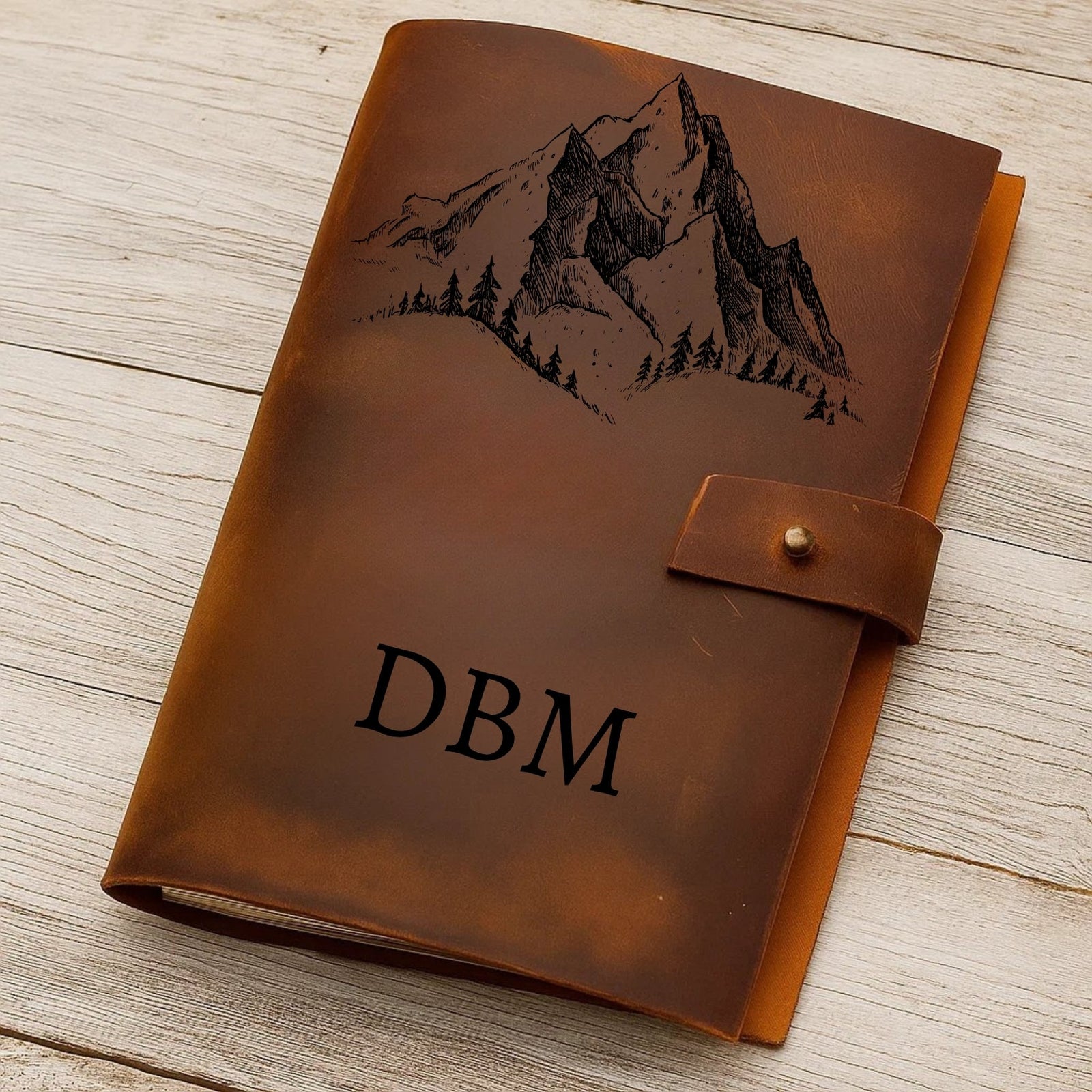 Brown leather journal with mountain engraving and 'DBM' initials on a wooden surface