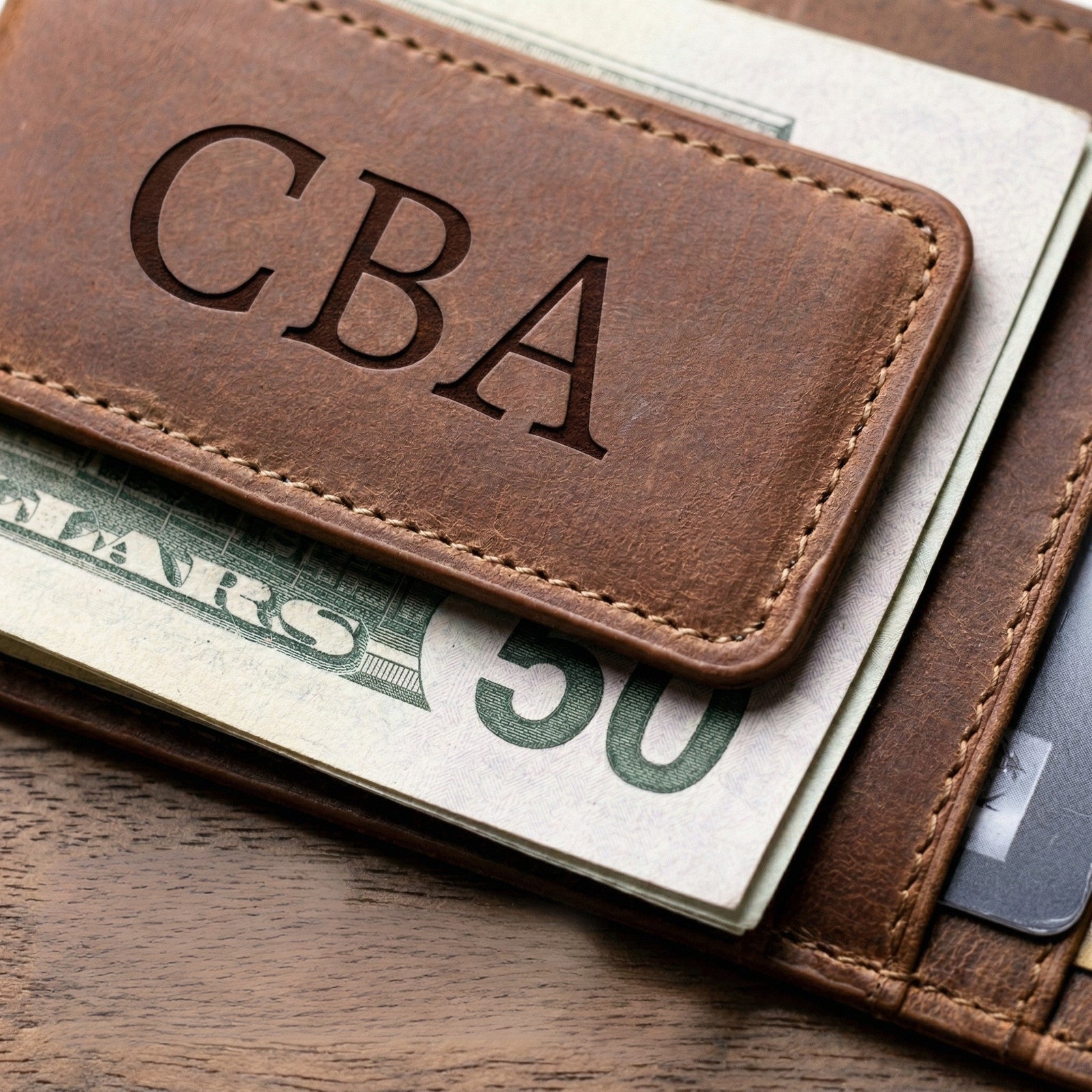 Brown leather wallet with 'CBA' engraving containing U.S. currency on a wooden surface