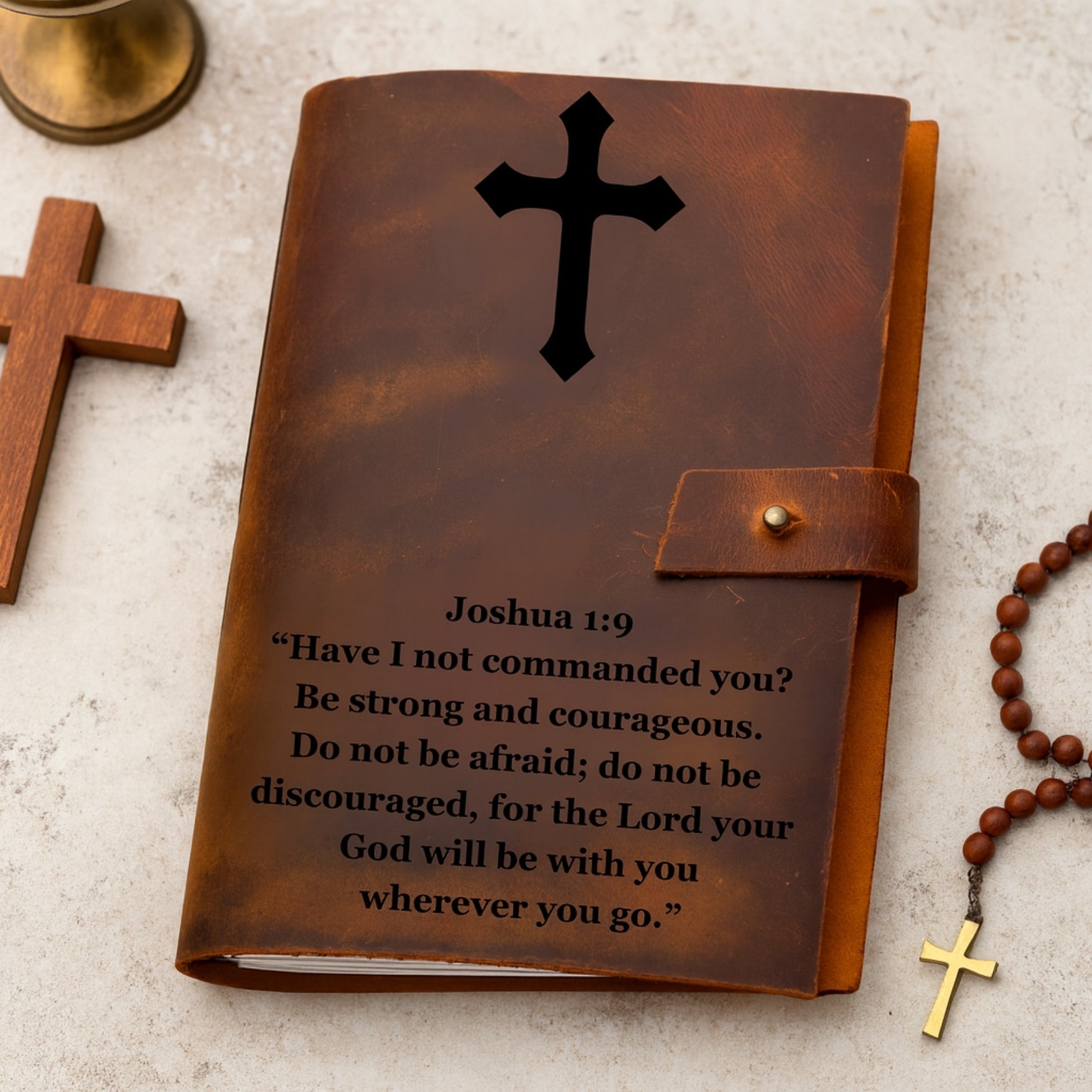 Brown leather journal with cross design and Joshua 1:9 verse on a light gray surface.